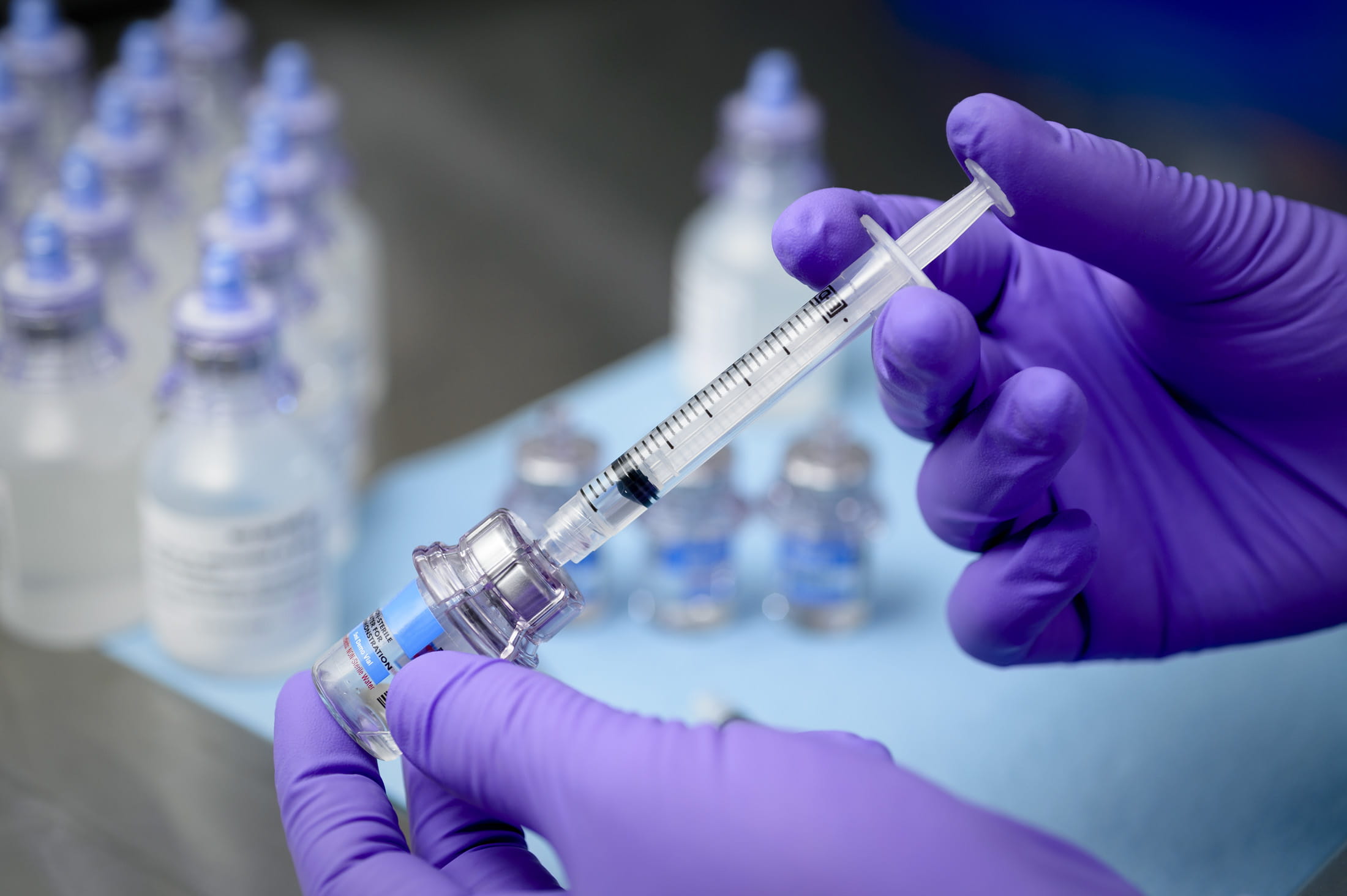Gloved hands drawing liquid from a vial with a syringe and vial adapter.