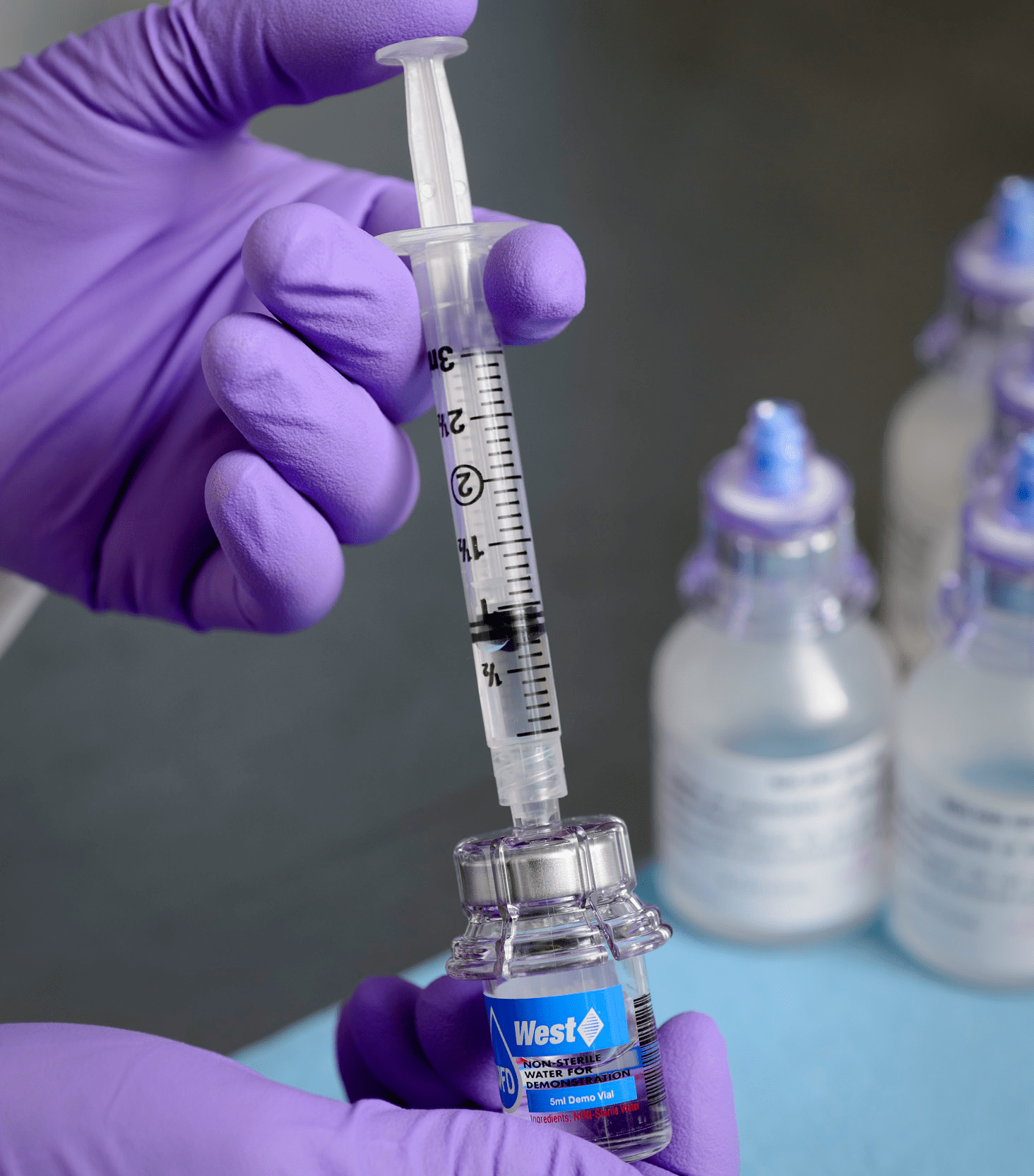 Gloved hand drawing liquid into a syringe from a vial fitted with a medical adapter.